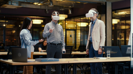 Multi-ethnic office workers testing VR headsets together, using tablet computer and wireless controllers, watching data, working in modern office of hi-tech company. Cyberspace digital technology