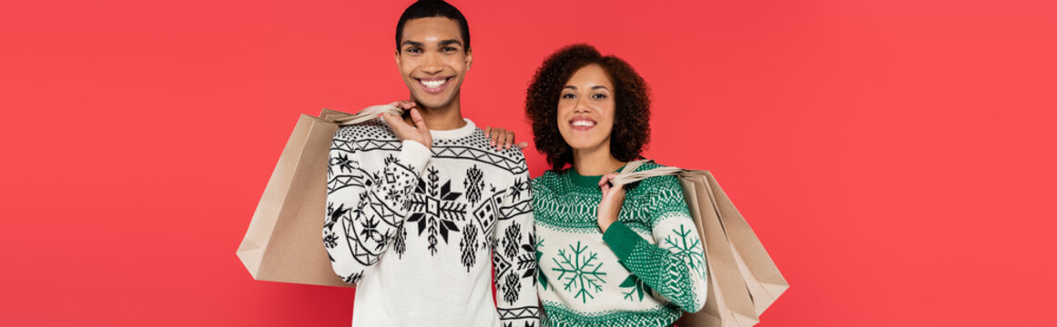 Pleased African American Couple In Sweaters With Winter Pattern Holding Shopping Bags Isolated On Red, Banner