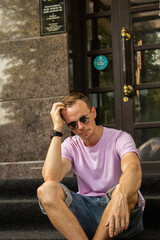 A handsome guy in denim shorts and a pink t-shirt poses beautifully in the city