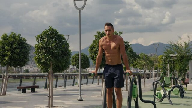 Man flexing abs on parallel bars in gym. Working on his core. Full length shot of a handsome and muscular young man working out on a dip bar outdoor