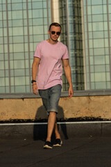 A handsome guy in denim shorts and a pink t-shirt poses beautifully in the city