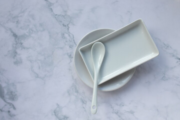 White tableware on marble table background. Saucer, rectangular plate, ceramic teaspoon. View from...