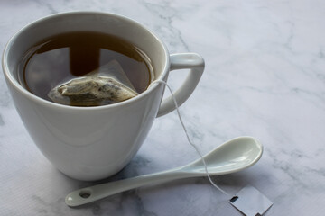 Cup of tea with tea bag and spoon on table. Green herbal tea in mug on white marble background. Top view. Traditional teatime. Teabag in cup 