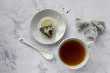 Cup of tea, tea bag on saucer and spoon on table. Green herbal tea in mug on white marble background. Top view. Traditional teatime. Teabag in cup 