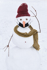 A confused snowman in a medical mask that is worn incorrectly. Puzzled snowman in a knitted hat and scarf on a background of snow.