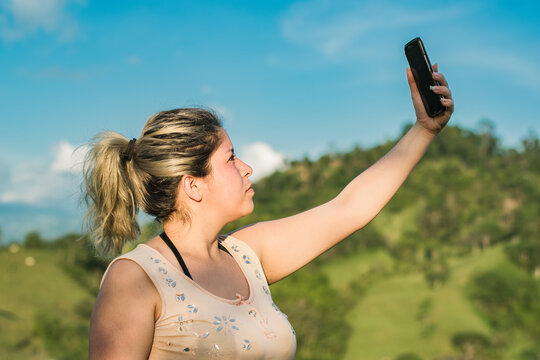 Beautiful Young Latin Woman On Top Of A Colombian Mountain Raising Her Hand To Look For A Signal For Her Mobile Phone. Girl Looking For Satellite Reception On Top Of A Hill. Satellite Internet Concept