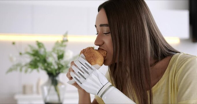 Girl using prosthetic arm while eating at home, woman with disability holds croissant in artificial prosthetic limb bionic hand