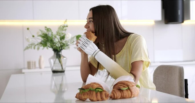 Girl using prosthetic arm while eating at home, woman with disability holds croissant in artificial prosthetic limb bionic hand