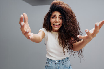 Wide angle view portrait of scared shocked screaming young Latin curly woman in white t-shirt looking and spreading hands at camera, expressing shock emotion, standing over gray studio background