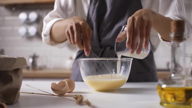 Female hands of a cook pouring milk into an egg yolk for making batter for baking pancakes, whipping liquid with kitchen metal whisk cooking breakfast
