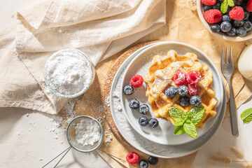 Traditional Belgian waffles stacked with fresh raspberries and blueberries sprinkled with powdered sugar on the kitchen counter