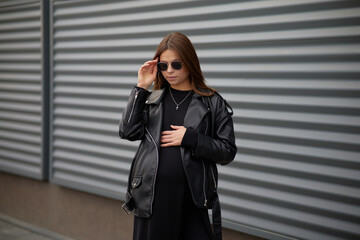 Stylish beautiful pregnant girl walks along a city street.Wearing a black leather jacket, a black long dress and white high boots and black sunglasses.