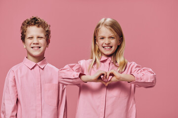 cute, beautiful children are standing in pink clothes on a pink background and the girl shows her heart with her hands, smiling broadly and closing her eyes