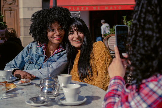 Young Interracial Female Friends, Latina And Haitian, Drinking Coffee On An Autumn Afternoon Outdoors, Take A Picture With A Cell Phone.