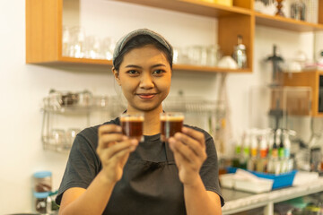 Young female barista welcome customer by holding two cups of expresso in front of her counter....