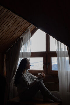Young woman sitting in cozy house and looking at window. Mountain background