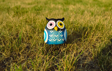 A toy of a surprised blue-colored owl with multi-colored eyes close-up stands in the middle of the grass in the light of the evening sun