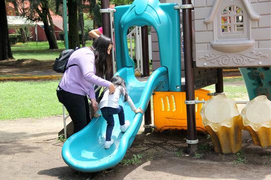 Single Divorced Latin Mom Plays With Her Daughter On The Slide In The Park Spending Quality Family Time Happily In Her Free Time On The Weekend Or Vacation