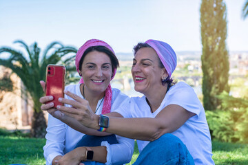 Two smiling young adult women with a pink headscarf taking a selfie. 2 warrior sisters laughing and happy after overcoming breast cancer. Breast Cancer Awareness Day concept.