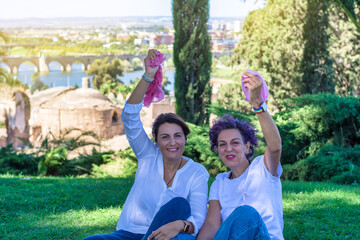 Breast Cancer Awareness Day concept. Two smiling women sitting on grass in an outdoor park holding up their pink scarves. Pink October