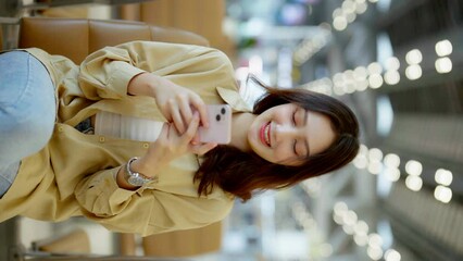 Vertical screen. Asian woman waiting for departure at the airport on vacation holiday. Asia female passenger using mobile smart phone and sitting in terminal hall while waiting for her flight - Powered by Adobe