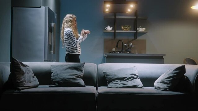 Woman enters kitchen, turns on lights with smart watch in her smart home IoT system. Female uses application on watch to control lightning scenarios in a living room, pouring wine. Internet of things.