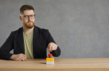 Serious man pushes big red button. Young hipster guy or businessman in glasses hits red alert button on table. Male entrepreneur sitting at desk presses activation button to get access to something