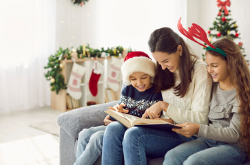 Happy young mother with small children celebrate New Year at home read book together. Smiling mom and little kids wear Santa hats enjoy literature on Christmas eve. Winter holiday concept.
