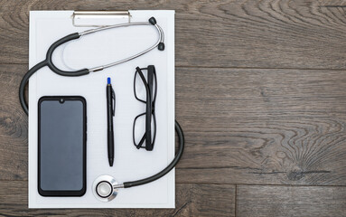 Top view on empty medical sheet, eyewear, smartphone, pen and stethoscope on wooden table with copy space. Place for text and medical health care concept