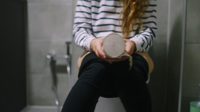 Low section of woman body sitting on toilet and suffers from diarrhea and stomachache. Female can't poop because of hemorrhoids piles. Strained girl holding and shaking toilet paper in hands.