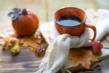 An orange mug in a scarf with hot tea, pumpkins, yellow dry maple leaves, a book on the windowsill, raindrops on the window - autumn mood