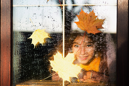 Child looks out of the window of the house outside, autumn weather, wet glass with drops after rain, yellow maple leaves stuck to the window. Autumn mood, home comfort