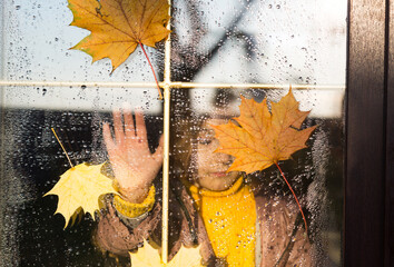 Child looks out of the window of the house outside, autumn weather, wet glass with drops after...