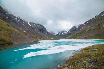 Obraz premium Atmospheric mountain landscape with frozen alpine lake and high snowy mountains. Awesome overcast scenery with icy mountain lake on background of snow mountains in low clouds. Scenic view to ice lake.