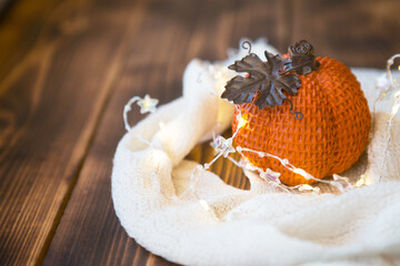 Orange pumpkin wrapped in a warm white plaid with lights of garlands on a wooden table with copyspace. Autumn mood, halloween, harvest, thanksgiving, home decor and cozy. Selective focus