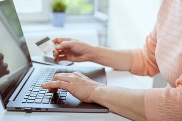 Mature woman works at home on a laptop. She is holding credit card in her hand. Closeup of hands, casual wear. Online shopping, work from home and freelance concept.