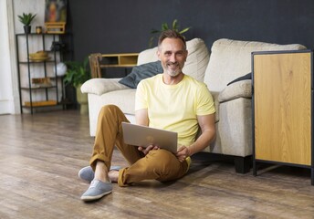 Portrait of handsome smiling man 45s wearing smart casual dress sitting on floor with laptop...