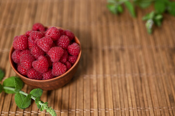 Fresh juicy organic raspberries in a clay bowl. Bamboo mat, green sprig of mint, space for text.