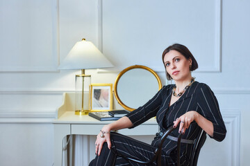 A stylish middle-aged woman in dark clothes is sitting on a chair in a retro-style room.