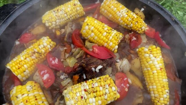 Corn with beef and vegetables. Shurpa in a cauldron. Tomato, red pepper and lamb soup cooked on hot coals. Hot beef hunting goulash or bograch soup with corn cobs, paprika, vegetables and spices.
