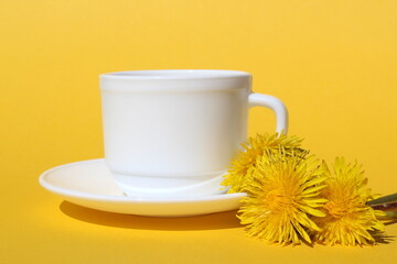 On a yellow background stands a white cup with a saucer and dandelion flowers.