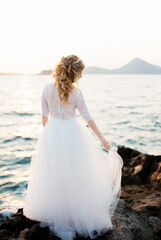 Fototapeta na wymiar Bride holds the hem of a layered dress on the rocks by the sea. Back view