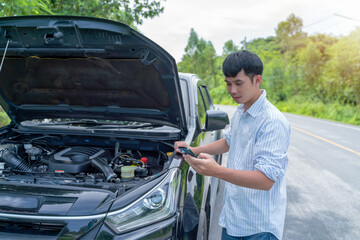 Handsome man inspecting a broken car looks at the engine under an open hood in the countryside and calls phone a mechanic.