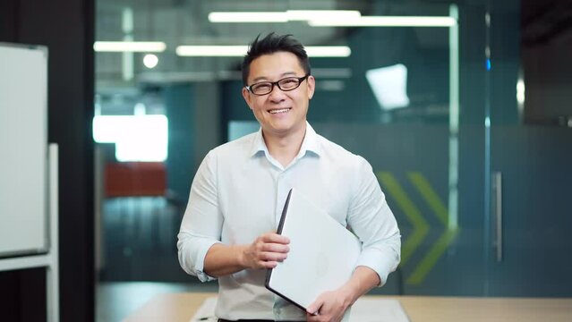 portrait of young asian man specialist, employee programmer or teacher standing in modern office or classroom at workplace. Happy male wearing glasses is looking at camera and smiling indoors