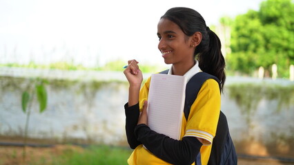 Smiling student girl wearing school backpack and holding exercise book. Portrait of happy asian young girl outside the primary school. Closeup face of smiling hispanic schoolgirl looking at camera.