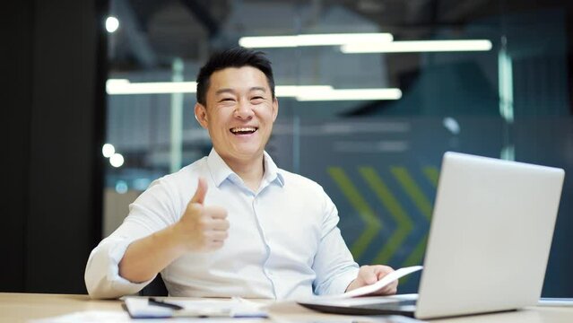 Portrait asian guy business men employee reading documents report paper project information letter bank funding notice rejoices feels happiness celebrating triumph sitting font of laptop looks camera