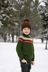 A smiling boy in a knitted hand-made sweater and hat in winter in a snowy forest