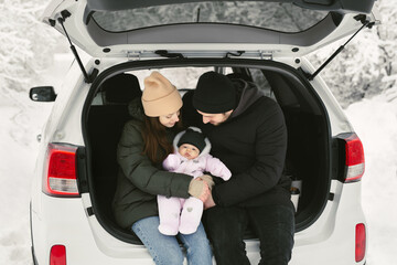 Young, happy family: a man, a woman and a baby are sitting in the trunk of a car in a winter snow-covered forest