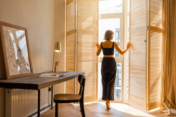 Woman opens window blinds letting the sun inside the room, spending good morning in sunny and cozy...