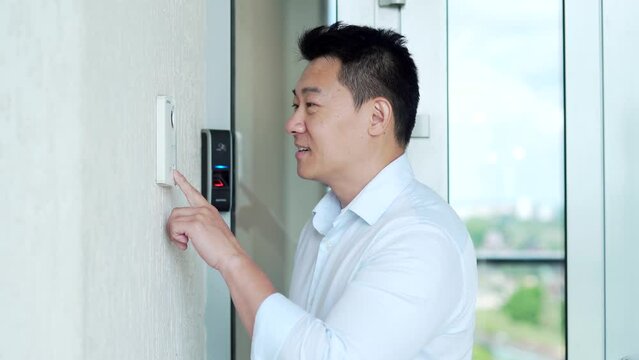 Asian man, calling rings the intercom of the house, pressing the button, waiting for the door to open. Male pushing the button and talking doorbell with camera near entrence electronic code lock